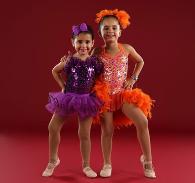 Two young girls in colorful dance costumes—one in purple sequins with a tutu and the other in orange sequins with feather accents—posing confidently against a red background.