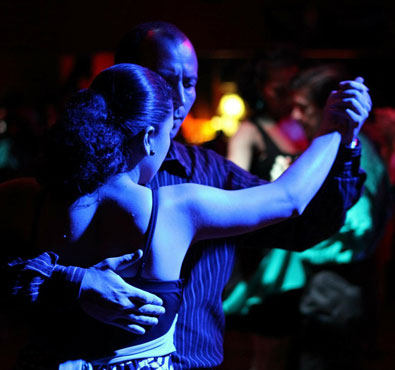 An instructor and student dancing closely together during a private lesson, illuminated by dramatic blue and warm ambient lighting.