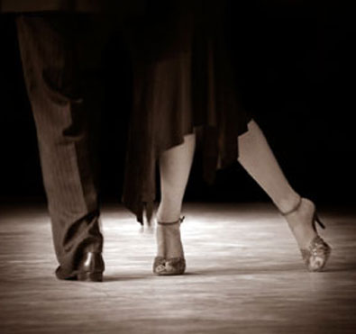 Close-up of a dance pair’s feet in motion—one in dress shoes and the other in heels—stepping together on a ballroom floor.