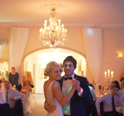 A bride and groom dancing together at their wedding reception under a chandelier, surrounded by guests in an elegant ballroom.