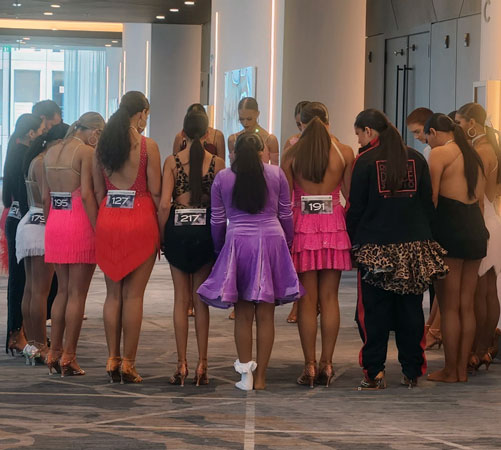 A group of young dancers in colorful ballroom costumes standing together in a circle backstage, preparing before a competition.